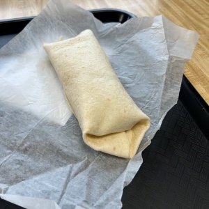A wrapped burrito sits on a sheet of wax paper atop a black tray, with a wooden table surface in the background.