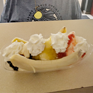A banana split dessert with three scoops of ice cream topped with whipped cream, chocolate, and strawberry sauce, served in a clear dish. A person in a gray shirt sits in the background.