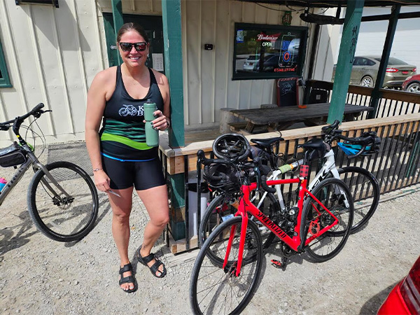 A woman in cycling gear stands smiling next to two parked bicycles outside Fiss' restaurant in Carlisle. She holds a green bottle and wears sunglasses, with sunny weather and parked cars in the background.