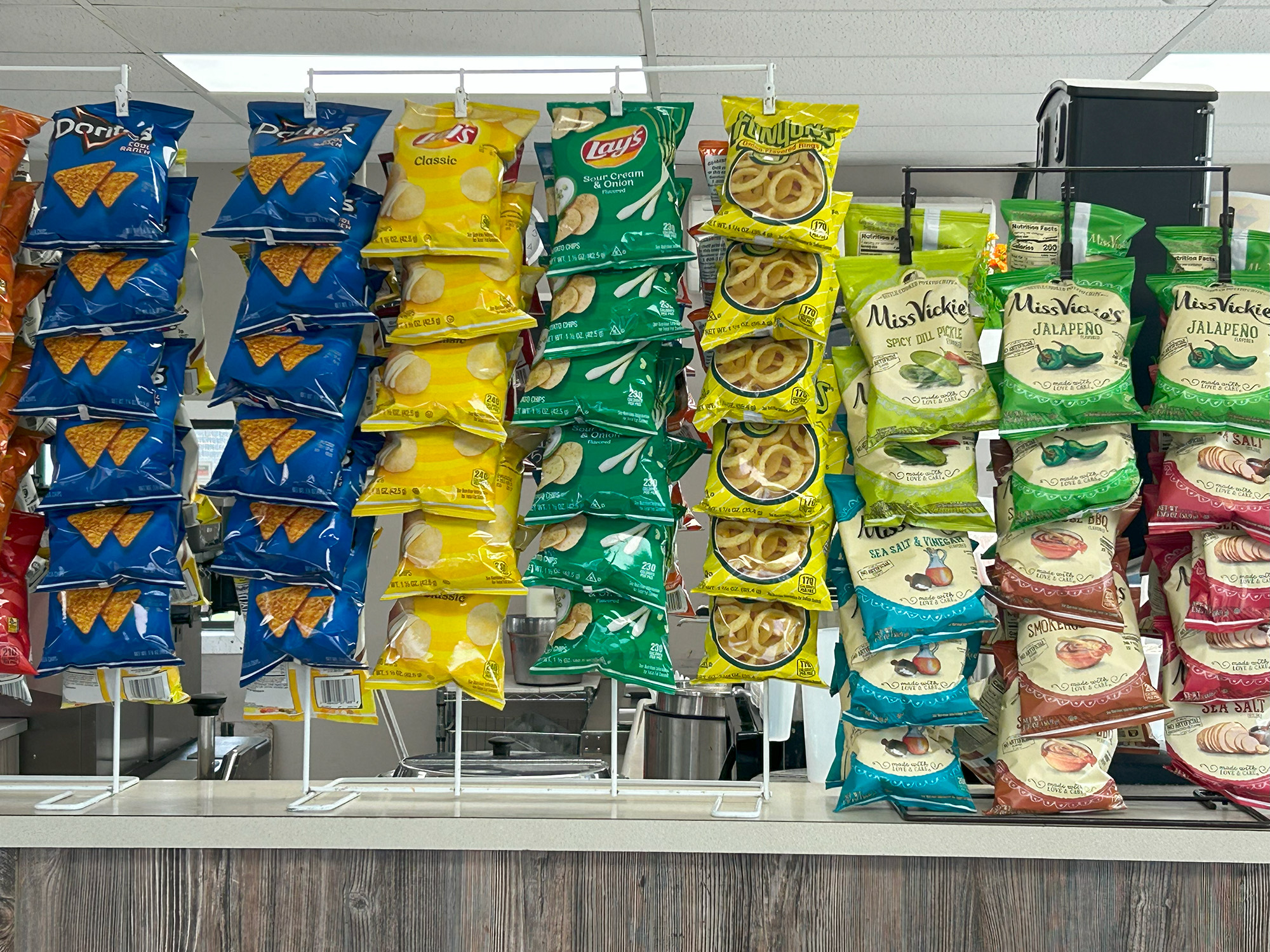 A display of assorted snack-size chip bags, including Doritos, Lay's Classic and Dill Pickle, and Miss Vickie's chips in flavors like Jalapeño, arranged on metal racks above a counter in a store.