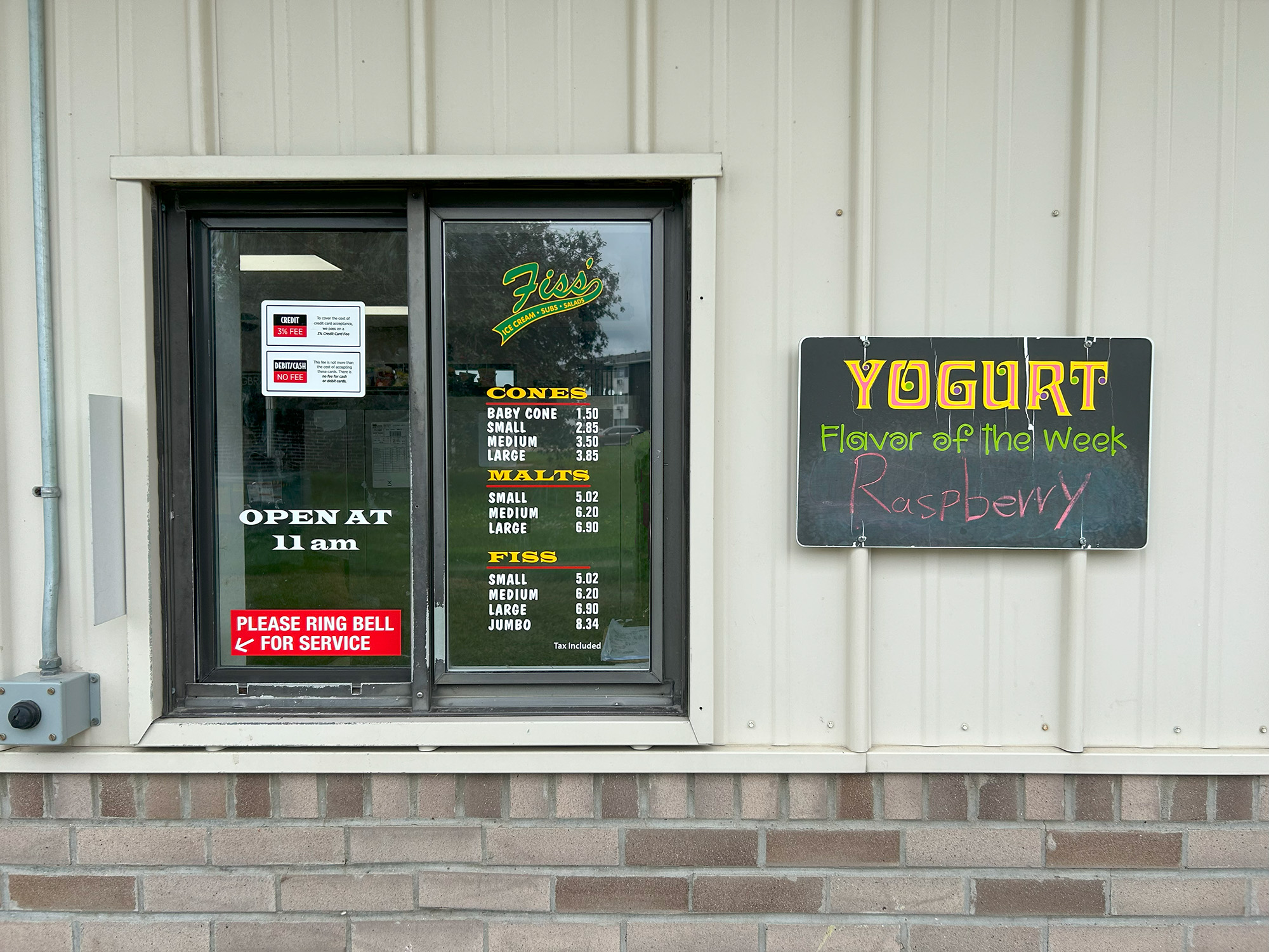 A walk-up service window with a menu listing cones, malts, and flis sizes and prices. Signs say “Open at 11 am” and "Please ring bell for service." A chalkboard reads, "Yogurt Flavor of the Week: Raspberry.