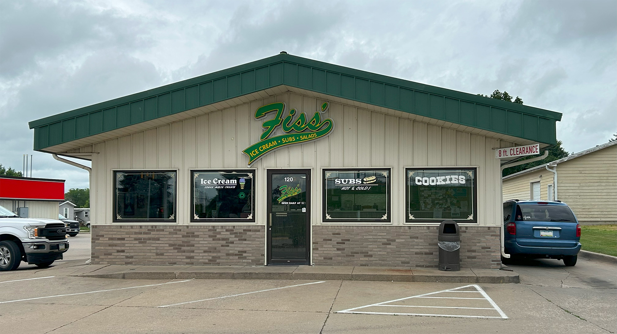 A small restaurant with a green roof named “Fiss’s Ice Cream, Subs, Salads.” Signs in the windows advertise ice cream, subs, and cookies. A blue minivan is parked outside. The sky is cloudy.