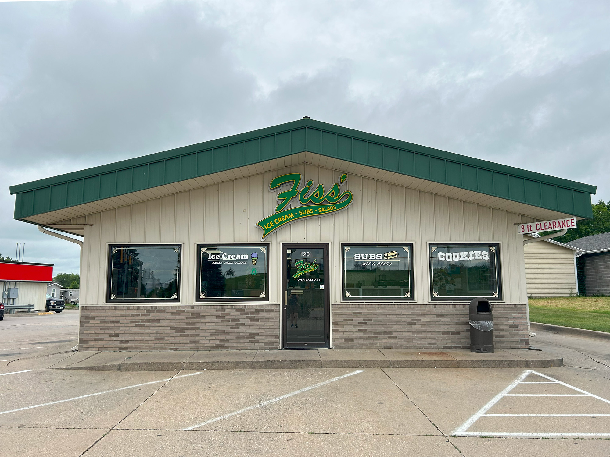 A small restaurant with "Fiss’s Ice Cream – Subs – Shakes" sign on the front. Windows display signs for ice cream, subs, and cookies. There’s a trash can and a handicap parking spot outside. The sky is overcast.