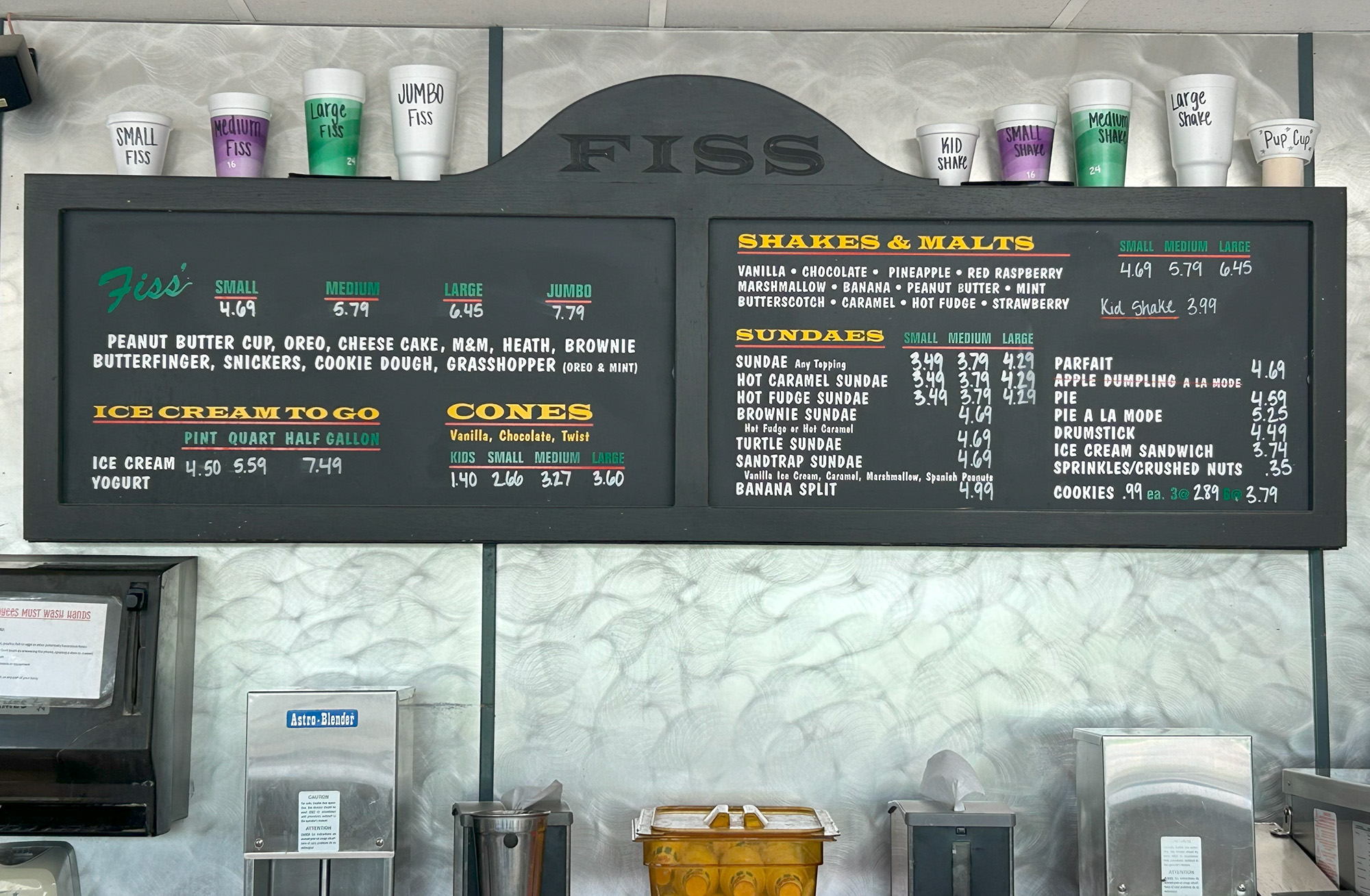 Menu boards at an ice cream shop display flavors, sundae options, cones, shakes, and prices. Branded cups line a shelf above the menu, and various toppings and containers are visible below the counters.