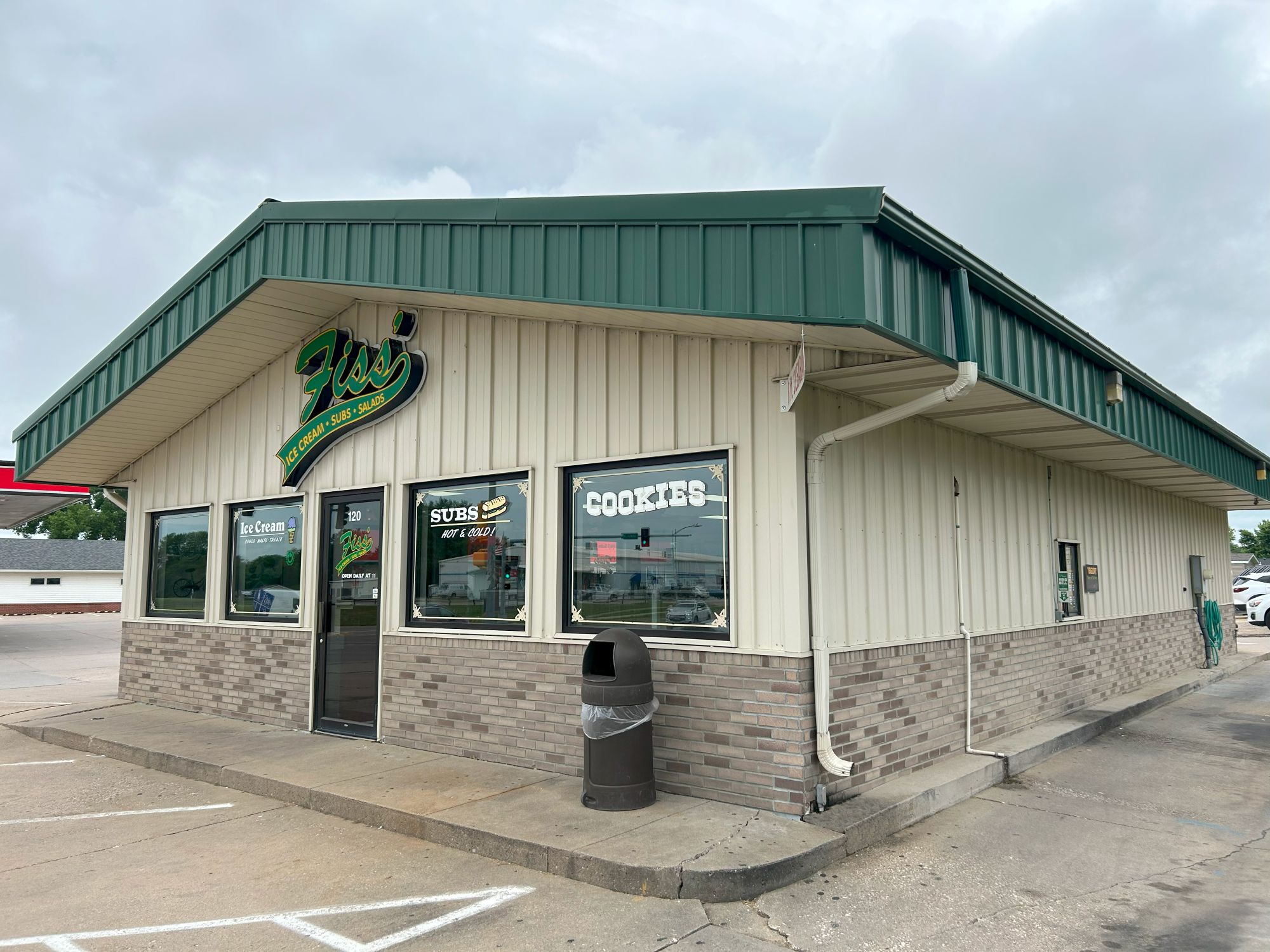 A small restaurant with a green metal roof and beige siding. Signs in the windows advertise subs, cookies, and ice cream. A trash can sits near the entrance. The sky is overcast.