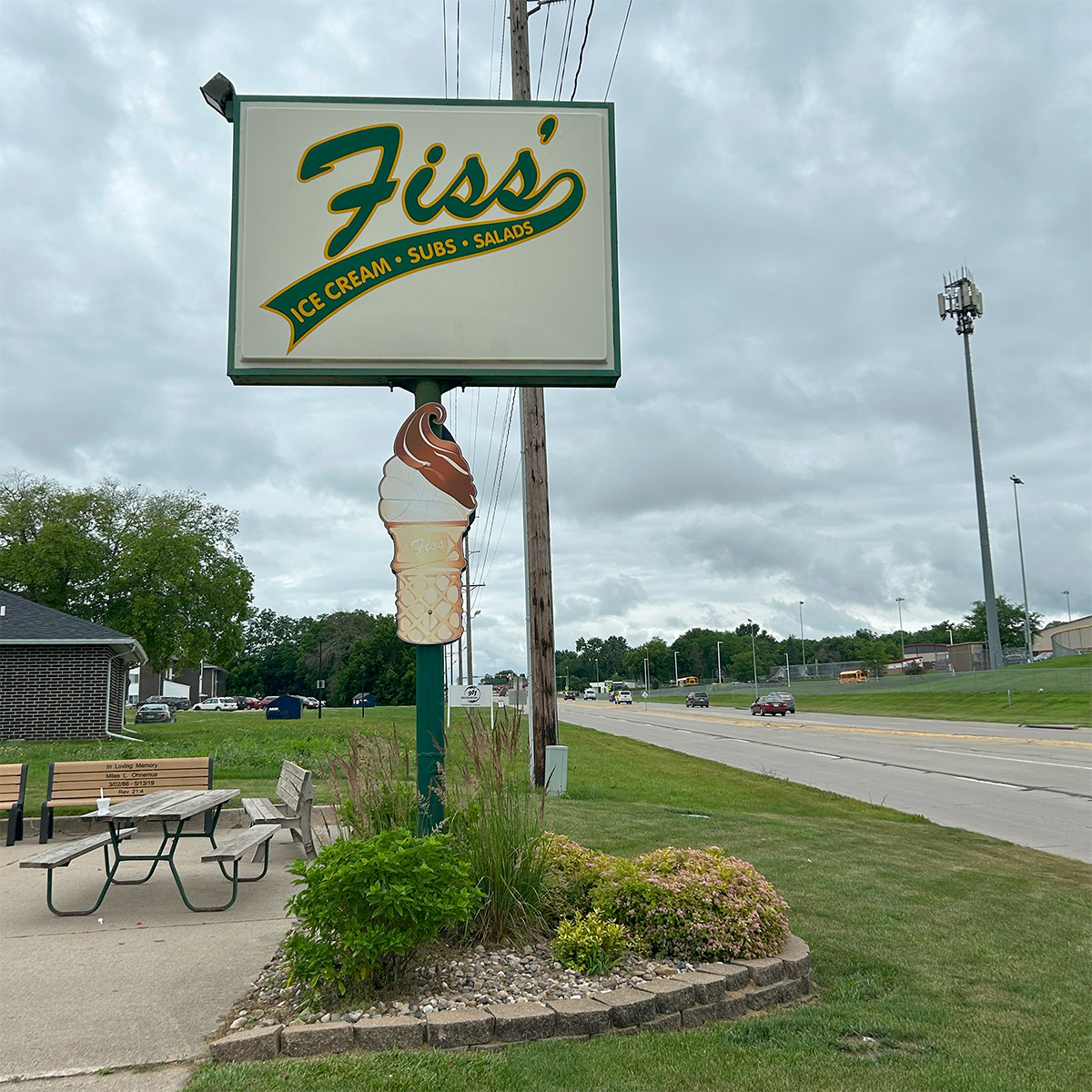 A large sign for Fiss's Ice Cream, Subs, and Salads stands by a roadside, featuring a graphic of a chocolate-dipped ice cream cone. There is a bench and table nearby, with cloudy skies overhead.