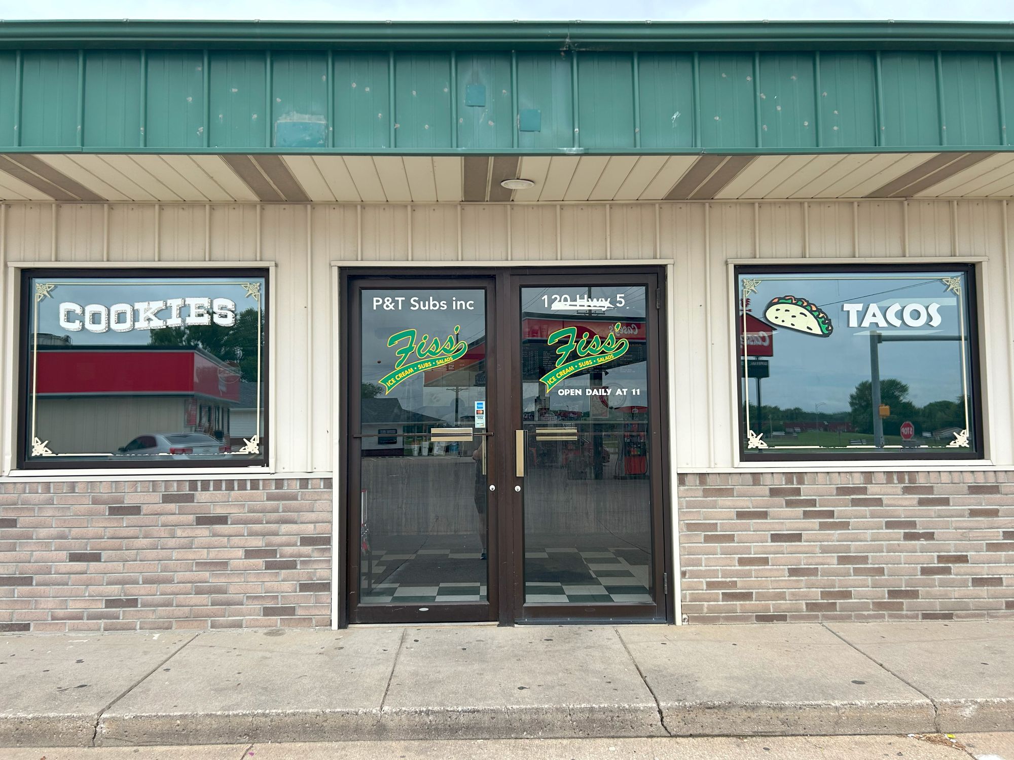 A storefront with glass doors and windows displays signs for "COOKIES" on the left, "TACOS" on the right, and "Ziss Subs" in the center. The building has a brick facade and a green metal roof trim.