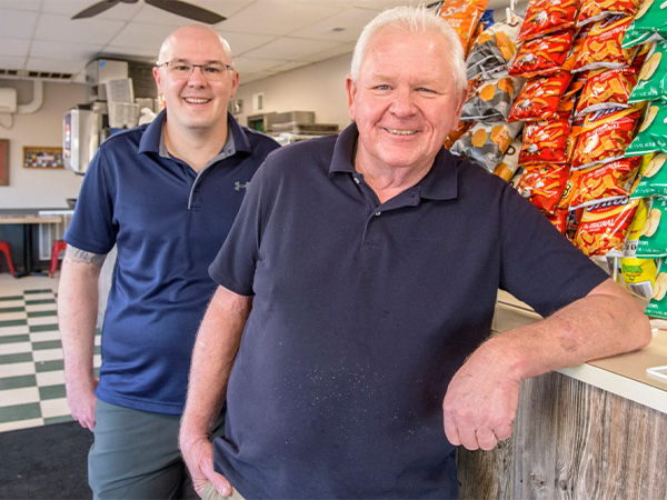 Tom and Matt Thiel smiling in dark polo shirts stand inside a Fiss' shop with a checkered floor; Tom leans on the counter near a display of assorted colorful chip bags.