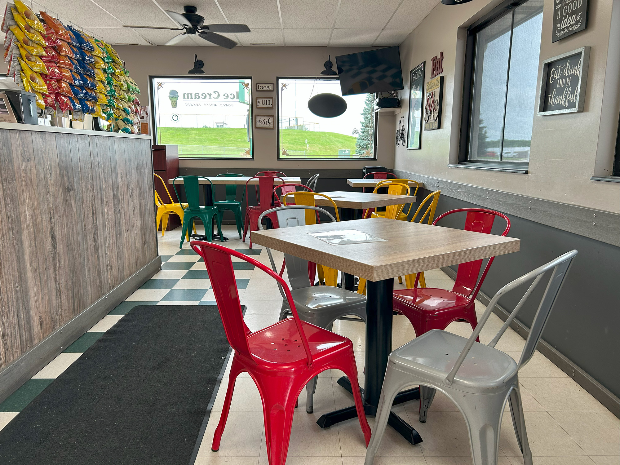 A small, casual eatery with colorful metal chairs around wooden tables, a wall display of bags of chips, checkered tile floor, and bright windows letting in natural light.