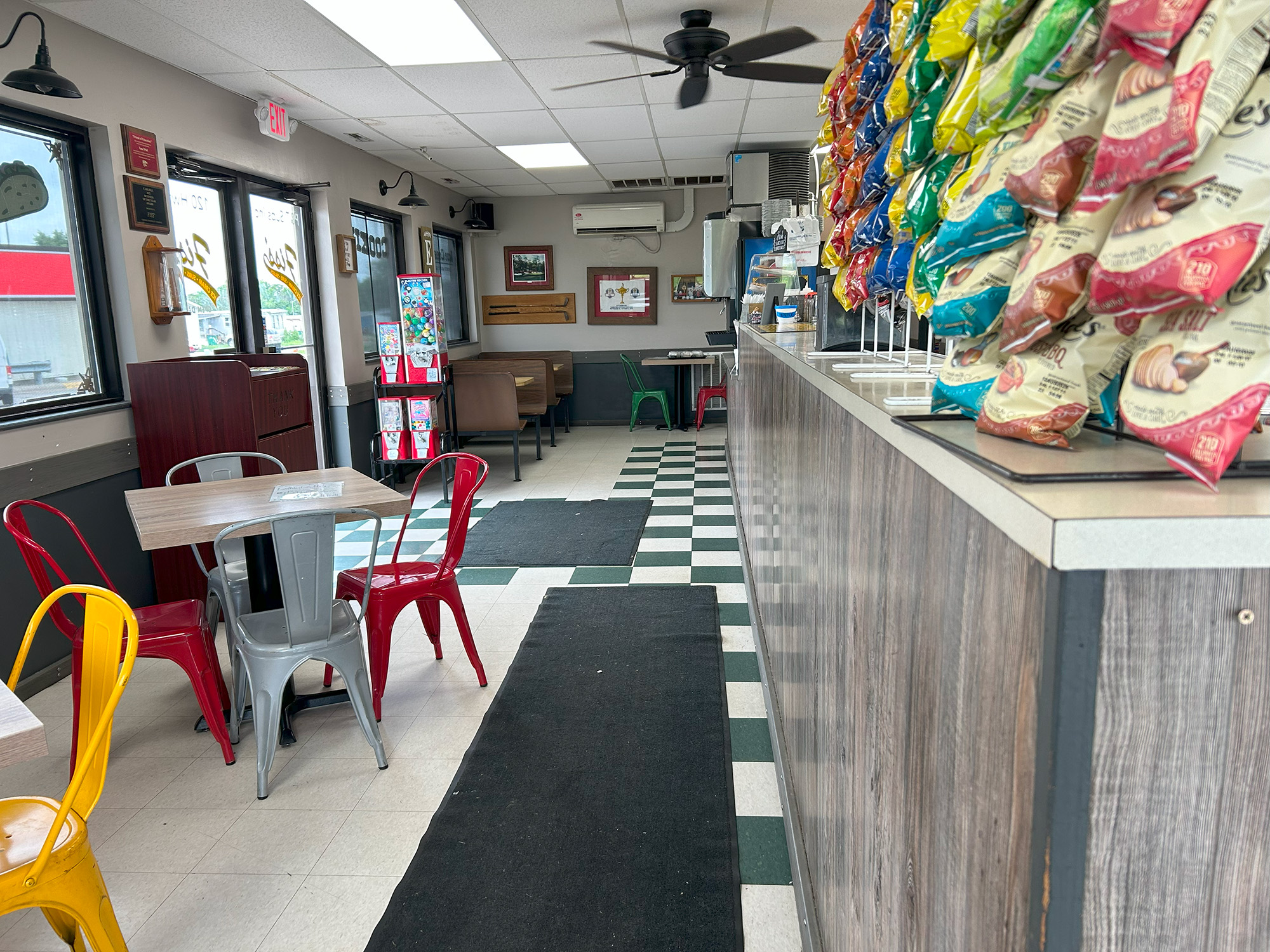 A view inside a Fiss' with checkered tile floor, red and yellow metal chairs around tables, a counter lined with colorful bags of chips, and daylight coming through large windows.