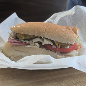 A sub sandwich with sliced tomato, lettuce, pickles, and cold cuts sits on a white paper plate, partially wrapped in white deli paper.
