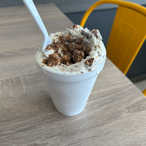 A styrofoam cup filled with cookies and cream ice cream, topped with chocolate cookie crumbles, sits on a light wooden table next to a bright yellow metal chair. A white plastic spoon is sticking out of the cup.