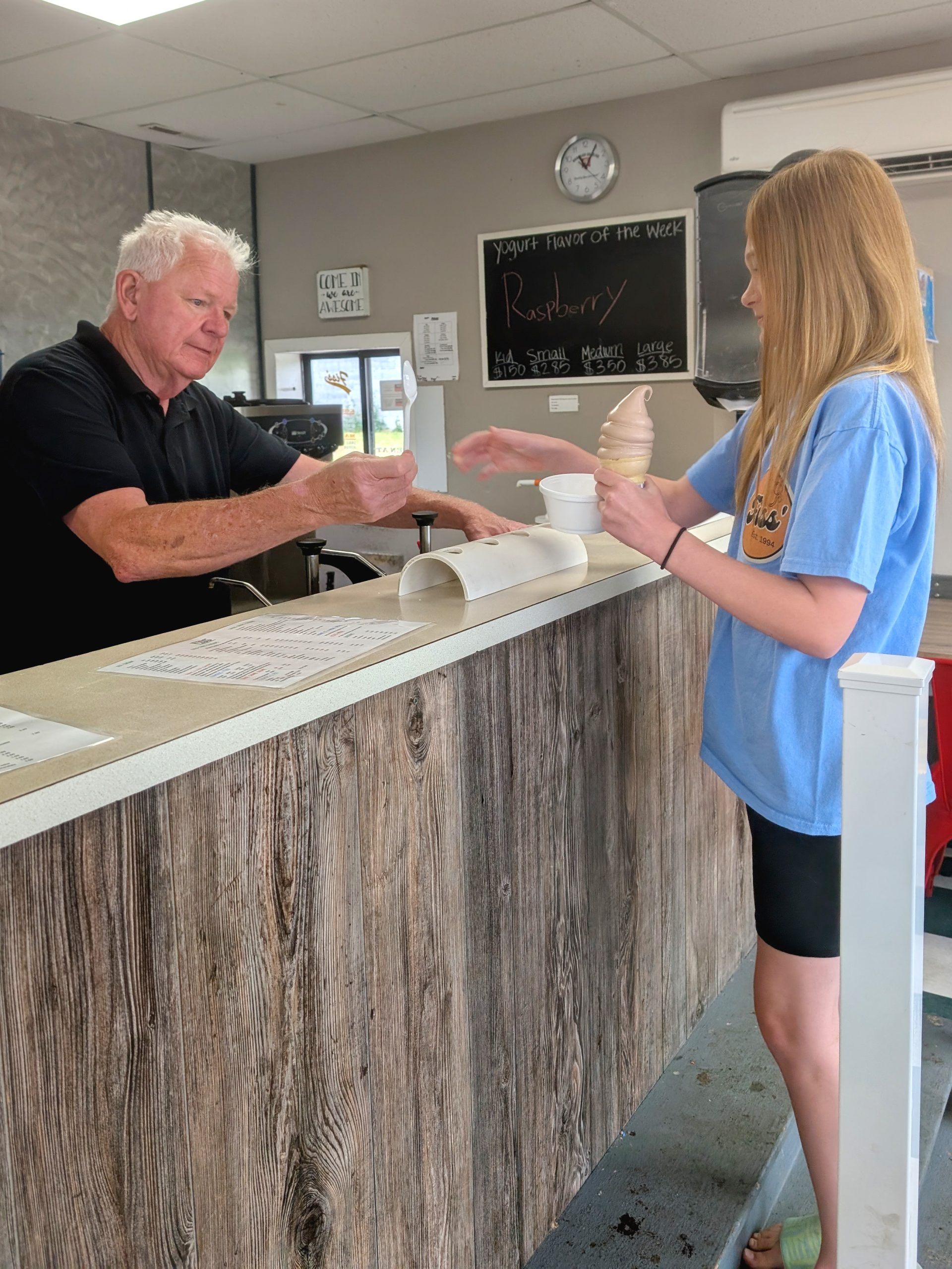 An older man hands change to a young woman holding a soft-serve ice cream cone at Fiss' ice cream counter. The shop has a rustic wooden counter and a chalkboard menu on the wall.